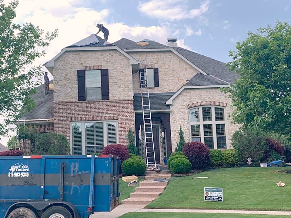 A roofer on a two-story stone and brick home in Allen, with a dump trailer and ladder in the drive