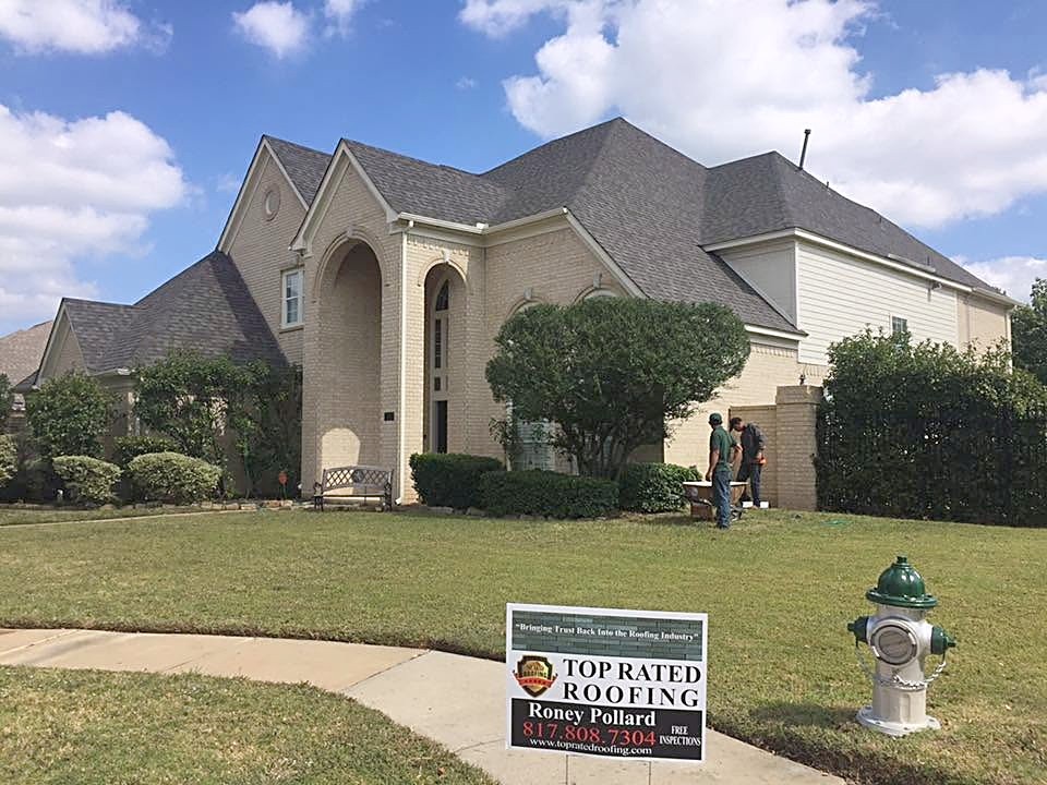 A beige two-story home with a new charcoal shingle roof, crew on the lawn and a Top Rated Roofing yard sign