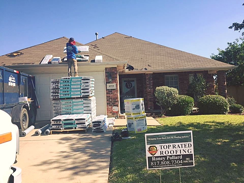 Pallets of shingles in a driveway while a roofer loads bundles onto a brick ranch roof