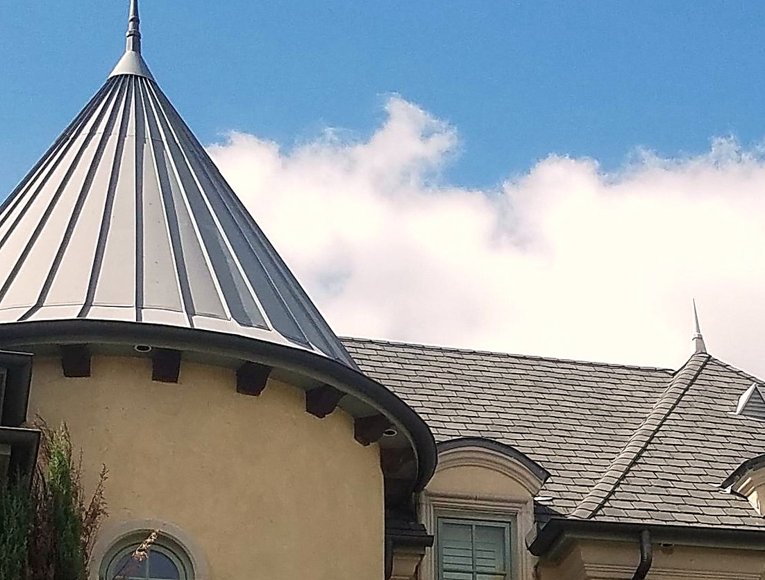 A standing seam metal turret roof beside slate-profile shingles on a stucco home