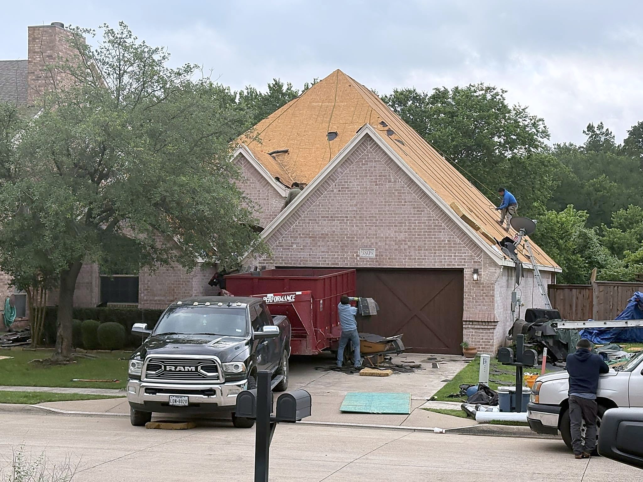 A two-story brick home mid tear-off, decking exposed, crew on the roof and a red dumpster in the drive