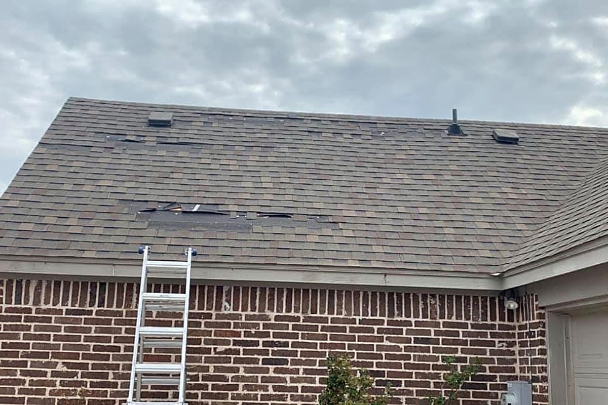 Missing shingles on the roof of a brick home after strong winds, a ladder against the wall